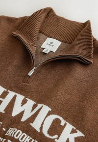Brauner Reißverschluss-Pullover mit geripptem Kragen und Bündchen, vorne mit weißem Text. Das Material wirkt gestrickt mit weicher, texturierter Oberfläche.