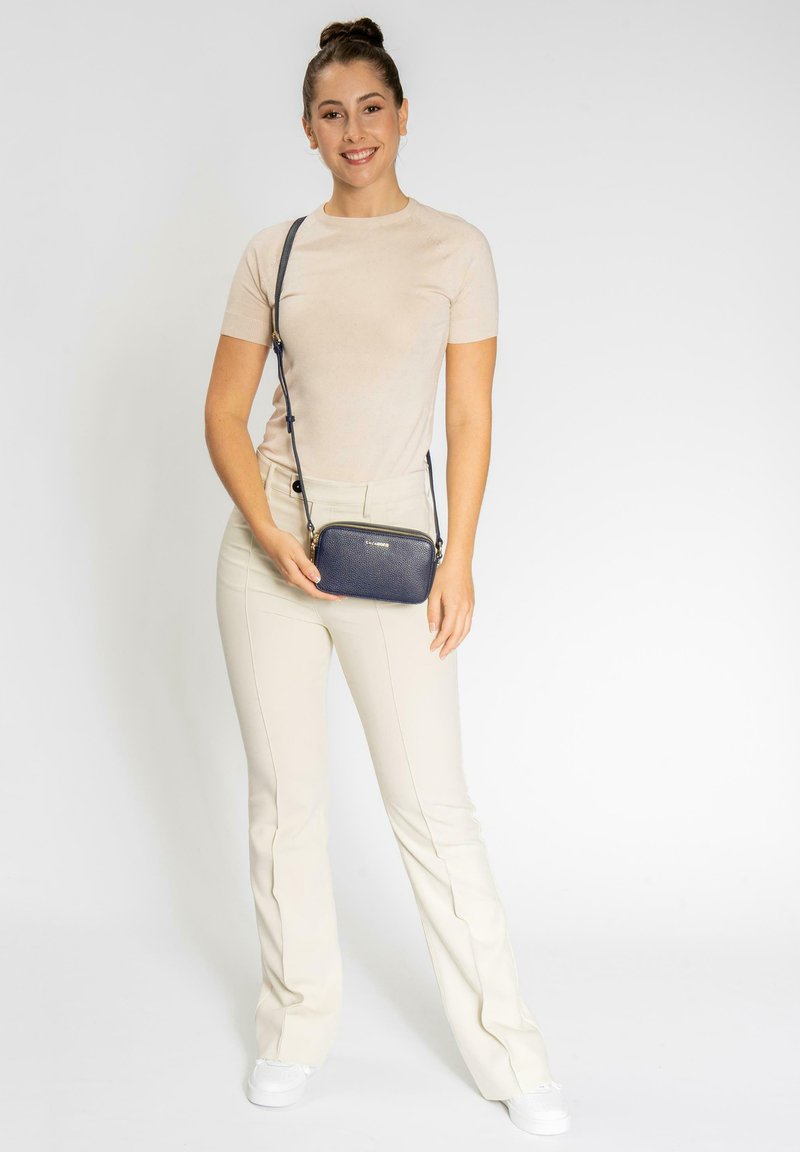 Borsa a tracolla testurizzata blu navy con chiusura a zip, tracolla regolabile e hardware in tonalità oro, posata su uno sfondo neutro.