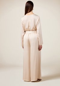 Beige, lange mouwen jumpsuit met een cinched taille en wijde benen. Glad, zijdezacht textuur met een subtiele glans en minimalistisch ontwerp.
