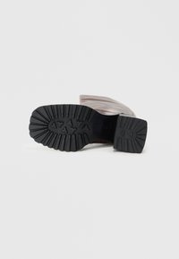 Une botte en cuir marron clair, couchée sur le côté, montrant une semelle en caoutchouc noire avec des motifs de semelle profonds sur un fond blanc.
