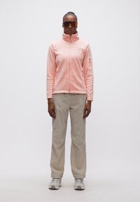 Γυναίκα που φοράει ανοιχτό ροζ φλις ζακέτα με φερμουάρ, ανοιχτό μπεζ παντελόνι cargo, άσπρα αθλητικά παπούτσια και μεγάλα αντανακλαστικά γυαλιά ηλίου, στέκεται μπροστά σε ένα απλό φόντο.