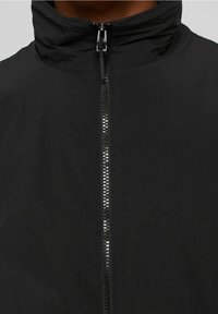 Giacca nera caratterizzata da un alto colletto e una zip riflettente. Il materiale sembra liscio con una finitura opaca, progettato per un look aderente.