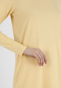 Oikea käsi lepää hellästi lanteen lähellä vaaleankeltaisessa pitkähihaisessa paidassa, jonka kangas on pehmeä.