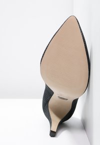 Chaussure noire à talons hauts avec un bout pointu et une surface texturée. Dispose d'une semelle beige lisse avec des rainures horizontales et un talon effilé.