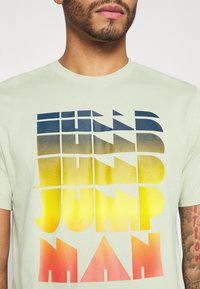 Camiseta de algodón verde claro con texto en bloques multicolor, que va del azul oscuro al amarillo y rojo, con cuello redondo y mangas cortas.