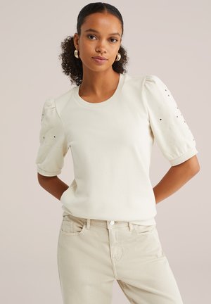 Top corto a maniche corte di colore crema chiaro, con maniche gonfie e dettagli ricamati, abbinato a jeans beige chiaro, che mostrano una texture liscia.