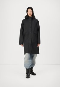 Svart regnjacka med huva, med tryckknappar, sidofickor och en mjuk textur. Kombinerad med överdimensionerade ljusblå byxor och svarta stövlar.