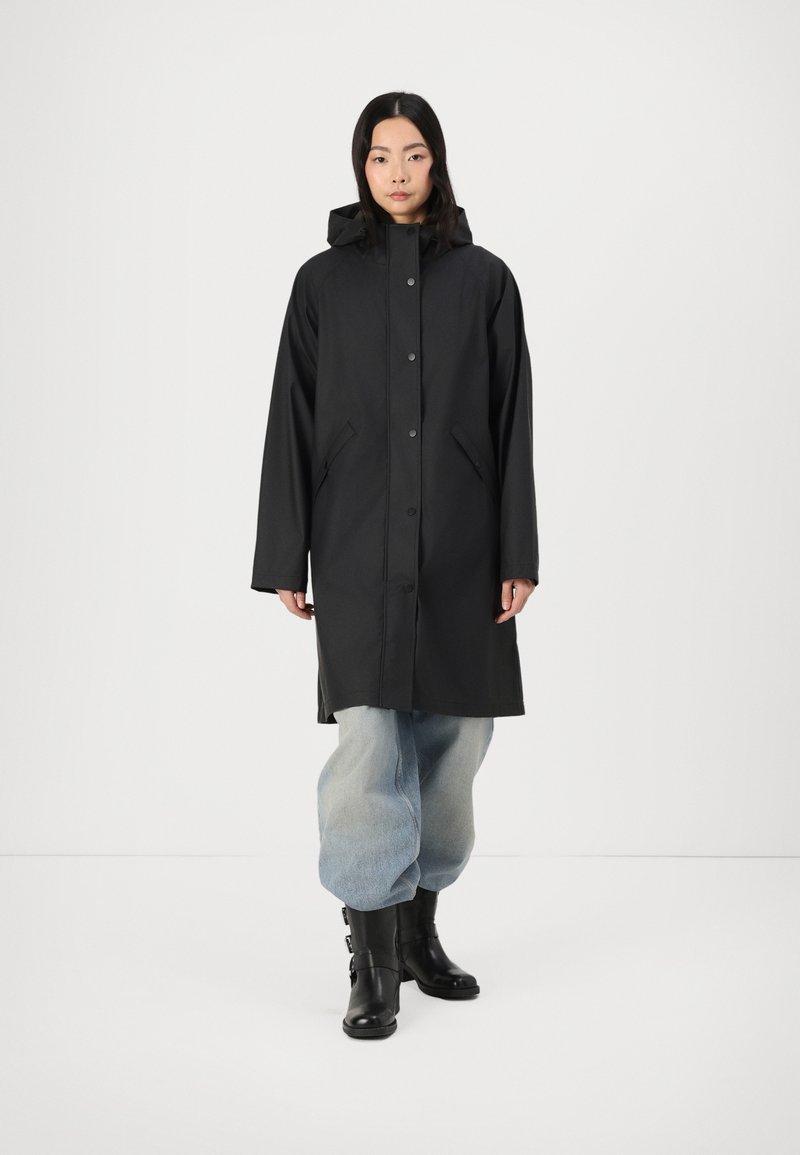 Svart regnjacka med huva, med tryckknappar, sidofickor och en mjuk textur. Kombinerad med överdimensionerade ljusblå byxor och svarta stövlar.