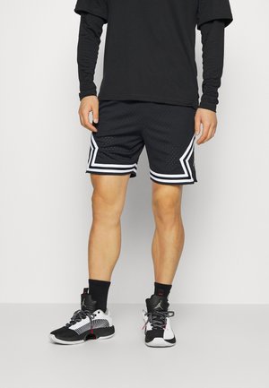 Mann trägt schwarze Sportshorts mit weißen geometrischen Streifen, schwarzes langärmeliges Shirt, schwarze Socken und schwarz-weiße Basketballschuhe.