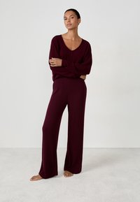 Donna in piedi a piedi nudi su un tappeto chiaro, che indossa pantaloni a gamba larga rigati bordeaux e un maglione a maniche lunghe con scollo a V coordinato.