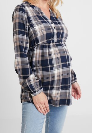 Femme enceinte portant un haut de maternité à carreaux bleu marine, beige et blanc avec des manches longues, et un jean bleu clair, sur un fond uni.