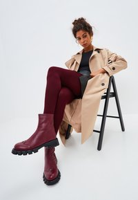 Bottines bourgogne avec semelle en caoutchouc noir texturé et couture notable, assorties à des leggings bordeaux et à un trench-coat beige.