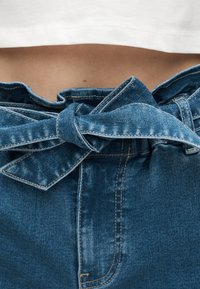 Κοντινό πλάνο δεμένης ζώνης από τζιν σε ψηλόμεσο μπλε παντελόνι με ορατή μέση και λευκό κοντό τοπ από πάνω.