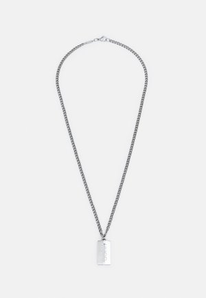 Collier chaîne argentée avec un pendentif rectangulaire gravé du mot « HUGO » sur un fond blanc uni.