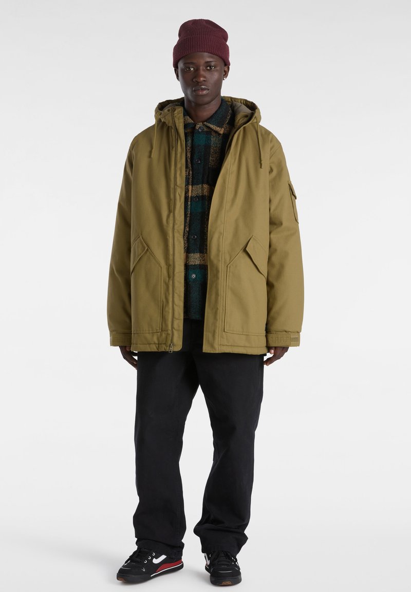 Chaqueta con capucha de color verde oliva en material duradero, sobre una camisa de franela de cuadros oscuros, combinada con pantalones negros y zapatos negros con detalles rojos.