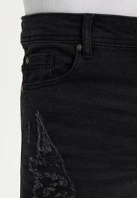 Pantalones vaqueros de mezclilla negra con diseño ajustado, que presentan detalles desgastados y costuras visibles cerca de los bolsillos. Detalles de botones metálicos presentes.