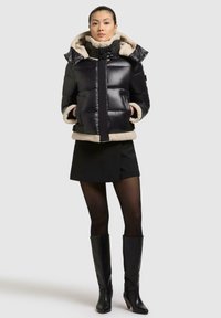 Schwarze Daunenjacke mit fellgefütterter Kapuze und Bündchen, ausgestattet mit einem Reißverschluss, einer aufgesetzten Tasche und einem verkürzten Design. Über einem schwarzen Miniröckchen getragen.