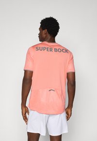 Camiseta deportiva rosa de manga corta, con un patrón texturizado, detalles en gris y "SUPER BOCK" en negrita en la parte posterior, combinada con pantalones cortos blancos.