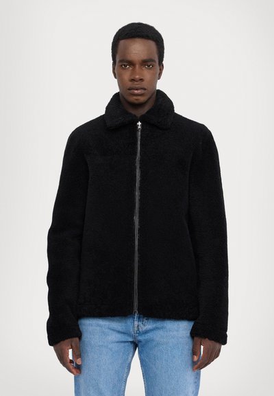 Veste polaire noire avec fermeture éclair sur le devant, col et manches longues. Texture douce, coupe décontractée, associée à un jean bleu clair. Sans motifs ni accents.