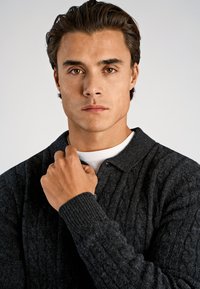 Ciemnoszary sweter z dzianiny warkoczowej z kołnierzykiem i prążkowanymi mankietami. Posiada teksturowany wzór i detal w postaci guzików. Pod spodem biała koszula.