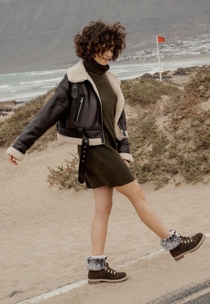 Mujer con cabello rizado vistiendo una chaqueta de borreguito, un vestido de punto oliva y botas con forro de piel caminando por un sendero de playa cerca del océano.