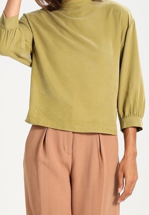 Femme portant un haut ample à manches longues vert olive avec manches bouffantes et col montant, assorti à un pantalon plissé marron taille haute.