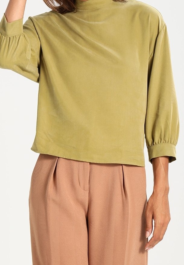 Femme portant un haut ample à manches longues vert olive avec manches bouffantes et col montant, assorti à un pantalon plissé marron taille haute.