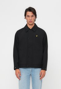 Veste noire avec un col, boutons sur le devant et deux poches latérales. Présence d'un logo d'aigle jaune sur la poitrine gauche. Texture de tissu lisse.