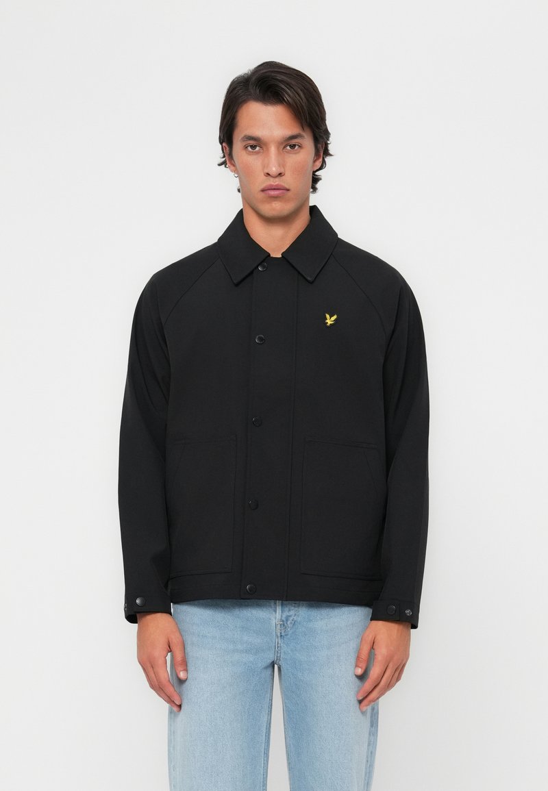 Veste noire avec un col, boutons sur le devant et deux poches latérales. Présence d'un logo d'aigle jaune sur la poitrine gauche. Texture de tissu lisse.