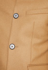 Hellbrauner Wollmix-Mantel mit zwei runden, schwarz akzentuierten Knöpfen, einer einzelnen Tasche und glatter Textur mit feinen Nähdetails.