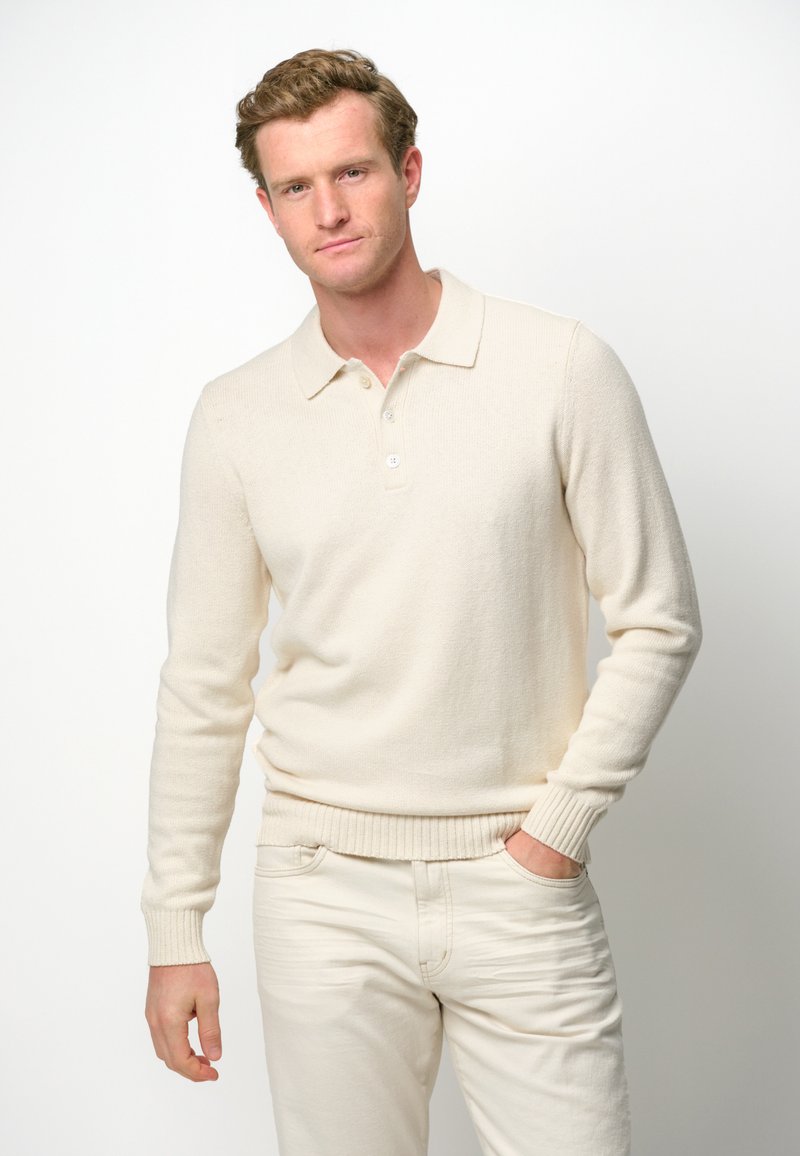 Cremefarbener Strickpullover mit kragenlosen Halsauschnitt, gerippten Manschetten und Saum; kombiniert mit hellen Hosen. Weiche Textur, lässiges Design.