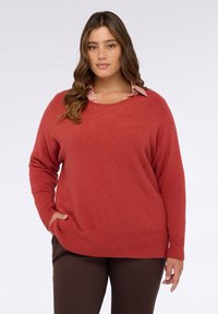 Maglione rosso oversize con scollo rotondo e orlo a coste. Tessuto morbido, maniche lunghe e tasche laterali. Indossato sopra una camicia rosa chiaro.