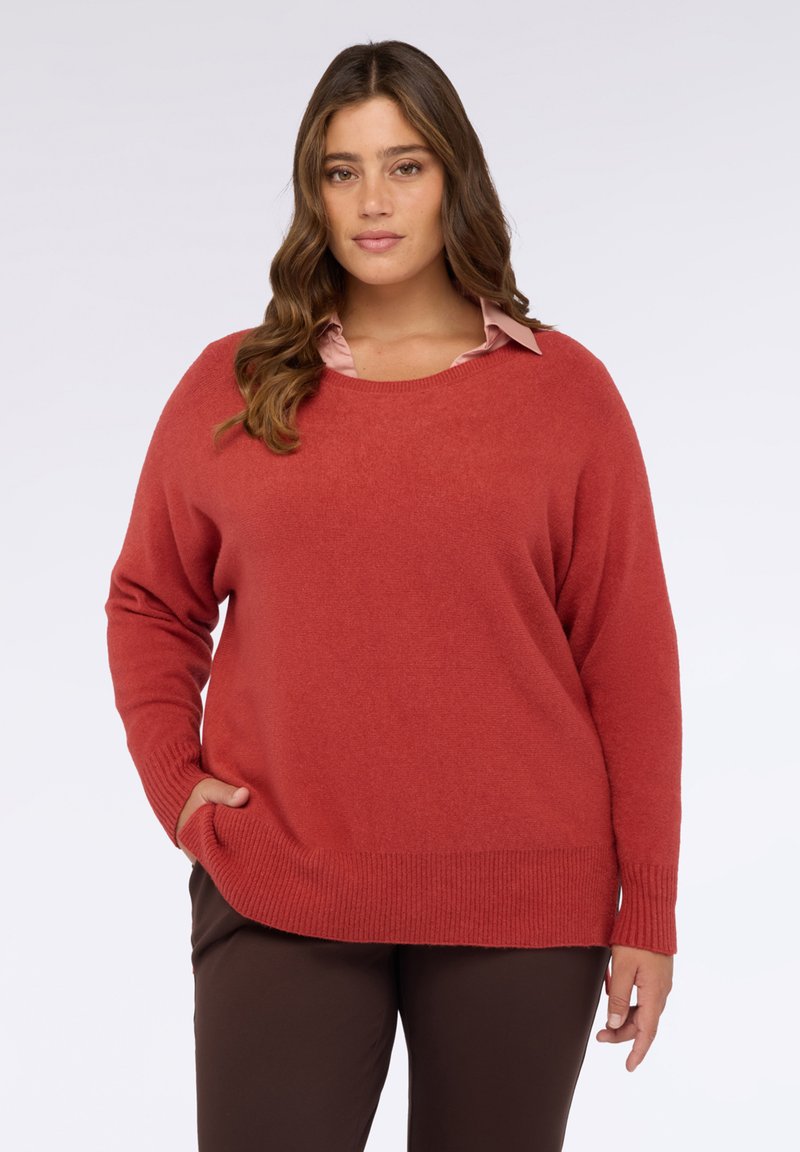 Maglione rosso oversize con scollo rotondo e orlo a coste. Tessuto morbido, maniche lunghe e tasche laterali. Indossato sopra una camicia rosa chiaro.