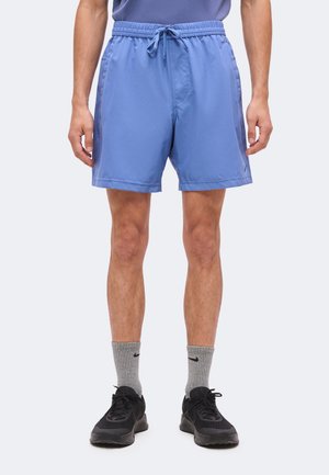 Homme portant un short de sport bleu, des chaussettes grises Nike et des chaussures de course noires, debout devant un fond uni.