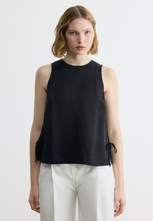 Młoda kobieta z krótkimi blond włosami, ubrana w czarny bezrękawnik z wiązaniami po bokach oraz białe spodnie, na jednolitym tle.