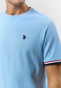 Svetlomodré bavlnené tričko s okrúhlym výstrihom, ktoré má malé čierne vyšívané logo a pruhované akcenty v červenej a námorníckej modrej farbe na rukávoch.