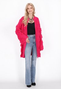 Cardigan deschis tricotat fuchsia cu franjuri texturati, asociat cu un top negru si blugi evazati de culoare bleu deschis, cu talie inalta. Pantofi negri completeaza look-ul.