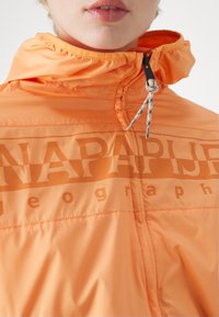 Pomarańczowa kurtka z wysokim kołnierzem ze sznurkiem, zamkiem błyskawicznym z przodu oraz dużym logo "NAPAPIJRI" na piersi. Gładki, lekki materiał.