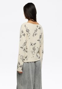 Pull crème avec des motifs floraux noirs, tissu texturé, coupe ample et épaules tombantes. Assorti à un pantalon large taille haute gris.