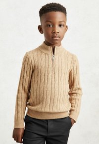 Pull en tricot beige à côtes avec un col zippé, présentant des motifs en câble texturés. Associé à un pantalon foncé, mettant en valeur une coupe ajustée.