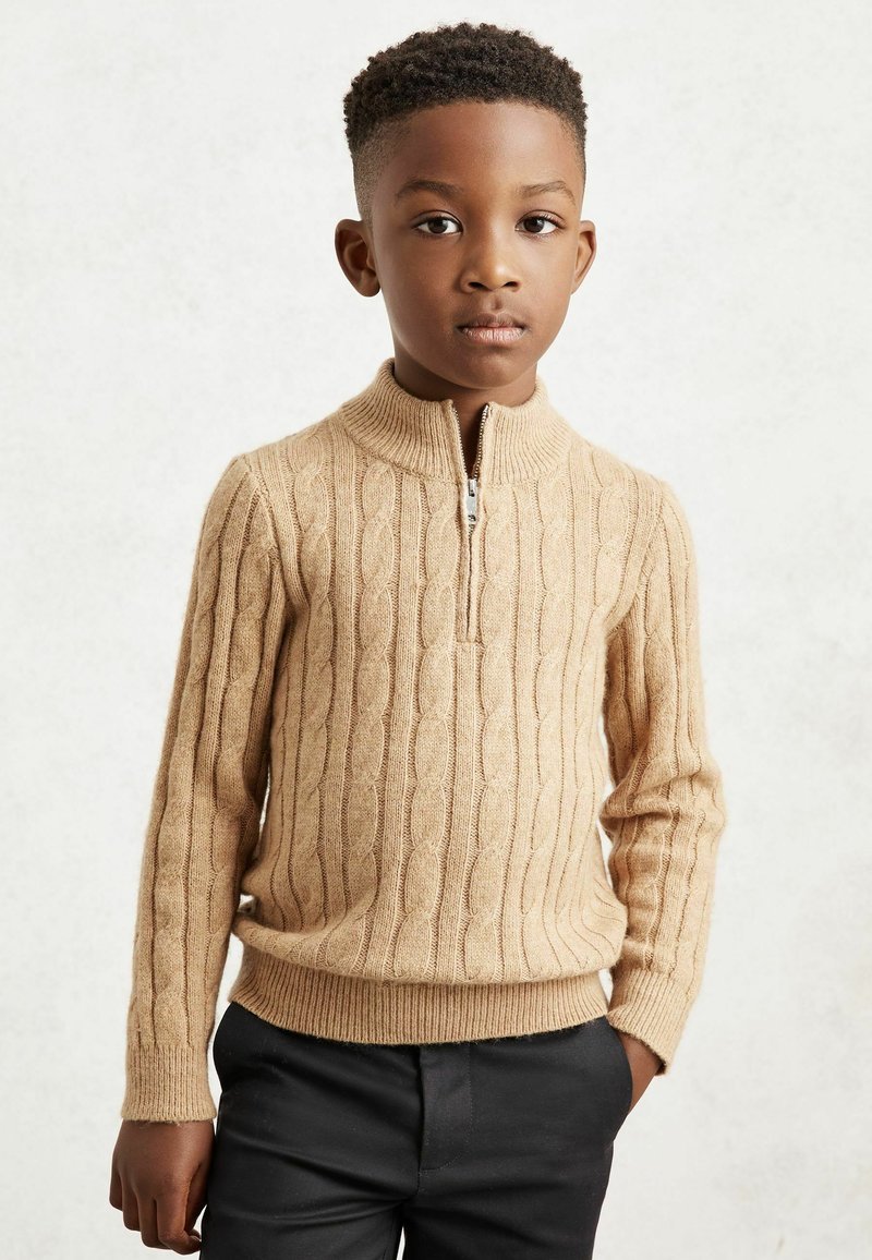 Pull en tricot beige à côtes avec un col zippé, présentant des motifs en câble texturés. Associé à un pantalon foncé, mettant en valeur une coupe ajustée.
