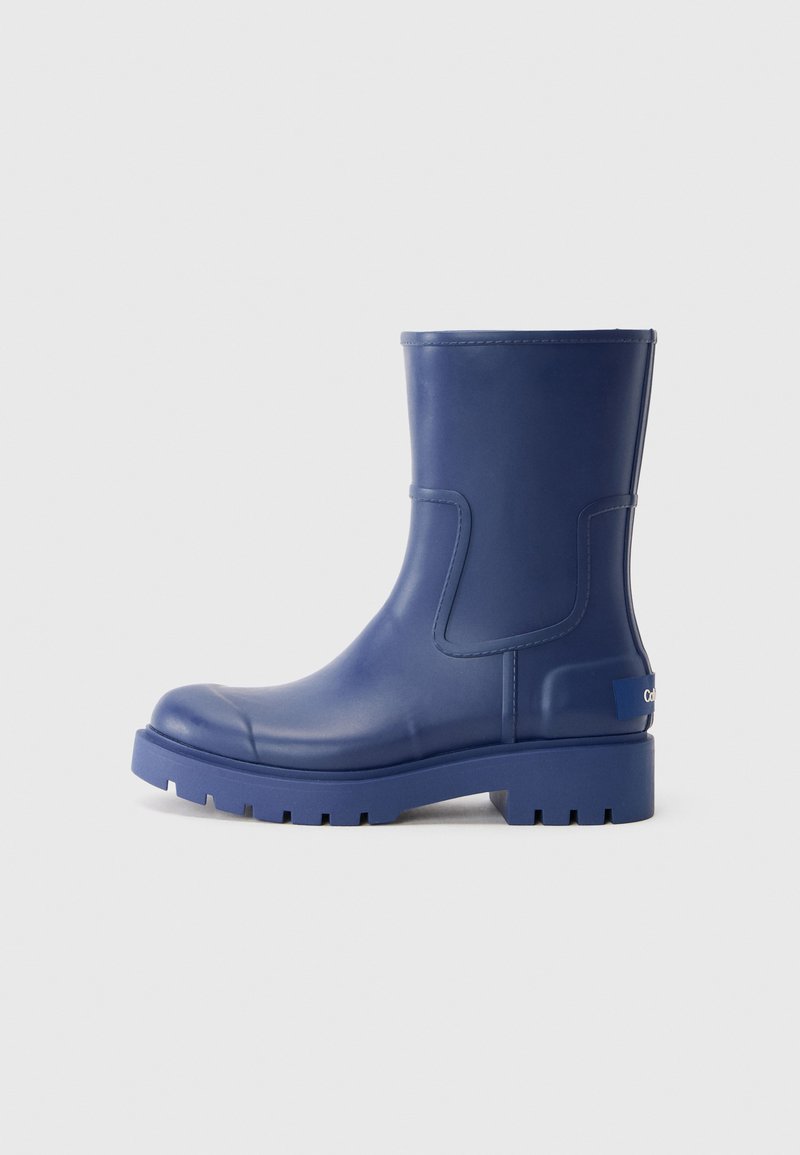 Bottes en caoutchouc bleues à la cheville avec un bout rond et une semelle texturée, présentant des coutures latérales surélevées et un accent de logo sur le talon.