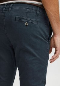 Tamno plave chinos hlače napravljene od mješavine pamuka; sadrže stražnji džep sa zatvaranjem na dugme i glatku teksturu. Jednostavan i prilagođen dizajn.