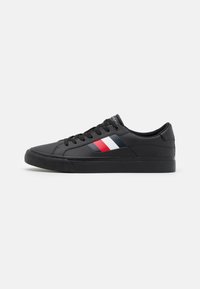 Zapatilla negra con suela de goma plana, parte superior sintética lisa y cierre de cordones. Presenta una franja diagonal en rojo, blanco y azul.
