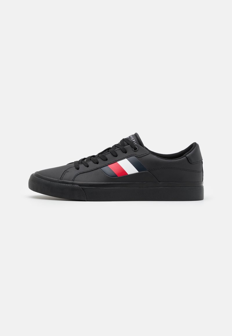 Zapatilla negra con suela de goma plana, parte superior sintética lisa y cierre de cordones. Presenta una franja diagonal en rojo, blanco y azul.