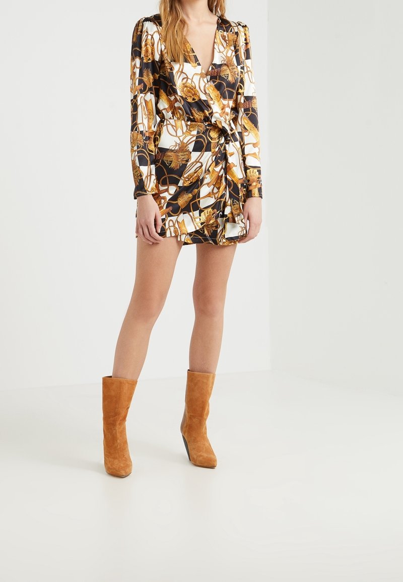 Femme portant une robe portefeuille à motifs noirs, blancs et dorés avec manches longues et bottines en daim beige sur un fond uni.