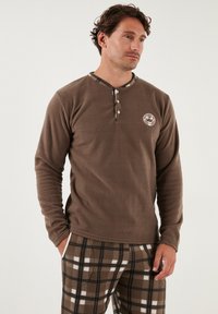 Haut long en polaire brun avec un patte de boutonnage à trois boutons et un logo. Associé à un pantalon à carreaux brun avec des carrés blancs et noirs.