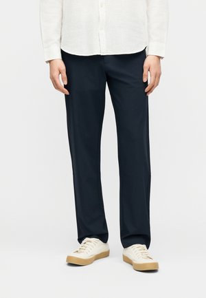 Άνδρας που φοράει μπλε σκούρες γραμμές παντελονιού, λευκό πουκάμισο με κουμπιά και λευκά αθλητικά με ταμπά σόλες, στέκεται μπροστά σε απλό φόντο.