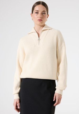 Pull en maille côtelée crème avec un col zippé mi-haute et un tirette en forme d'anneau argenté, associé à une jupe noire. Texture douce, coupe décontractée et manches longues.