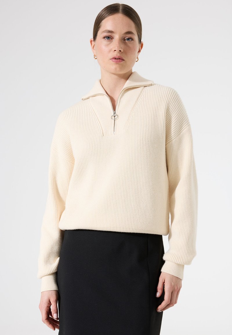 Suéter de punto acanalado crema con un cuello de media cremallera y un tirador plateado, combinado con una falda negra. Textura suave, corte relajado y mangas largas.